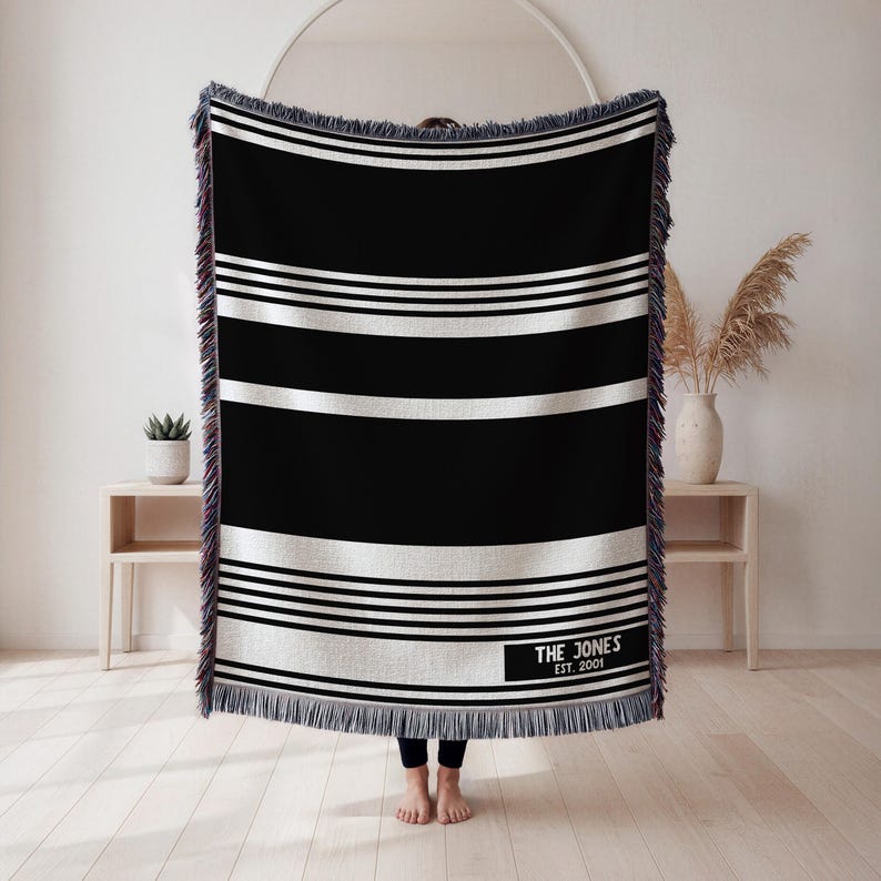 May include: A black and white striped throw blanket with a rectangular label that reads "THE JONES EST. 2001". The blanket has a fringed edge and is displayed in front of a white wall.