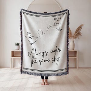 May include: A cream-colored woven throw blanket with fringed edges featuring a black line-art map of South Carolina labeled Sam and New York labeled Jessica, connected by a dashed line with hearts, and the text Always under the same sky.