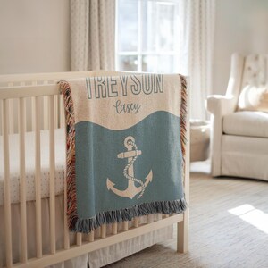 May include: A personalised baby blanket with the names "TREYSON" and "Casey" in a script font. The blanket features a blue anchor design and a fringed edge. The blanket is draped over a white cot.