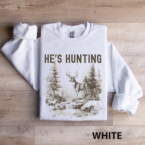 May include: White sweatshirt with the text "HE'S HUNTING" above a sepia-toned illustration of a deer in a forest scene. The word "WHITE" is printed at the bottom. The sweatshirt has long sleeves and a crew neck.