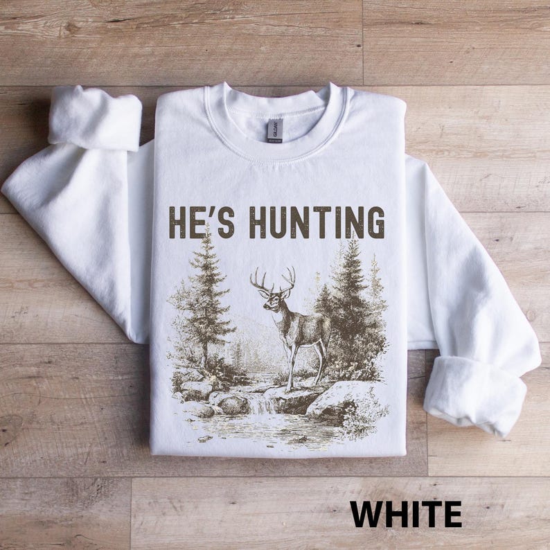 May include: White sweatshirt with the text "HE'S HUNTING" above a sepia-toned illustration of a deer in a forest scene. The word "WHITE" is printed at the bottom. The sweatshirt has long sleeves and a crew neck.