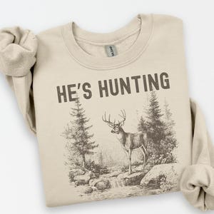 May include: A beige sweatshirt with the text "HE'S HUNTING" in dark brown. The graphic depicts a deer standing near a stream with trees in a forest setting. The sweatshirt has rolled-up sleeves.