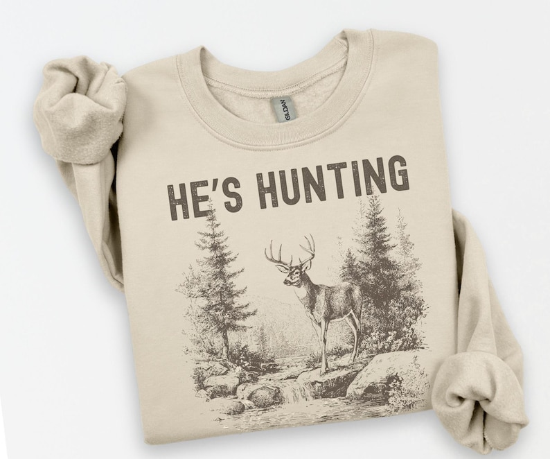 May include: A beige sweatshirt with the text "HE'S HUNTING" in dark brown. The graphic depicts a deer standing near a stream with trees in a forest setting. The sweatshirt has rolled-up sleeves.