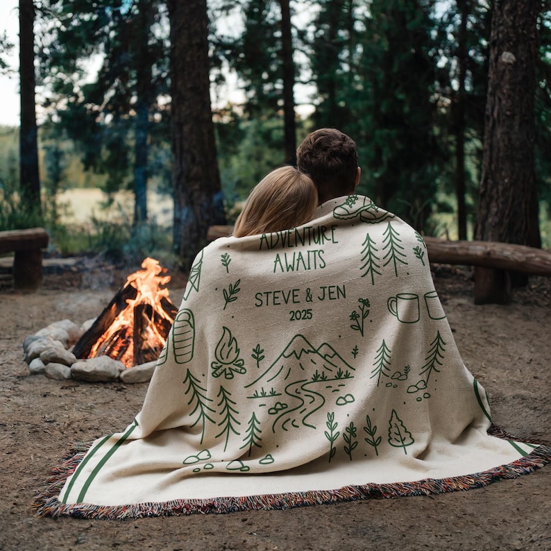 May include: A beige woven blanket with green trim and fringe, featuring a forest-themed design. The blanket has the words "ADVENTURE AWAITS" and the names "STEVE & JEN 2025" printed on it. The blanket is draped over two people.