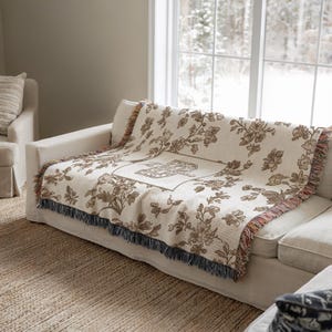 Op de afbeelding: Een cr&egrave;mekleurige sprei met een bloemenpatroon en franjes is over een witte bank gedrapeerd. De sprei heeft een centraal vierkant ontwerp en bruine bloemmotieven. De bank staat op een geweven vloerkleed bij een raam met uitzicht op een besneeuwde buitenomgeving.