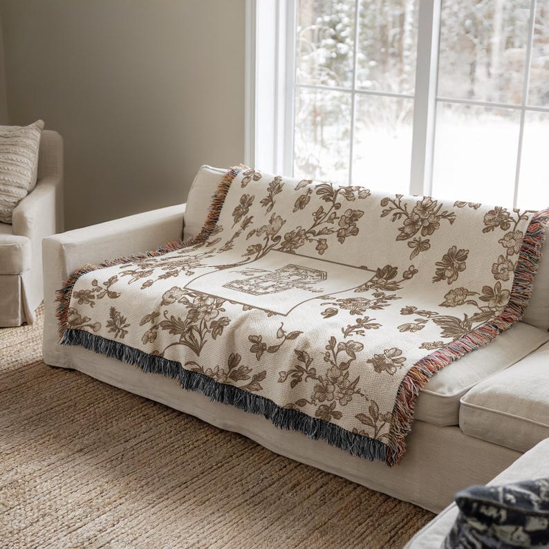 Op de afbeelding: Een cr&egrave;mekleurige sprei met een bloemenpatroon en franjes is over een witte bank gedrapeerd. De sprei heeft een centraal vierkant ontwerp en bruine bloemmotieven. De bank staat op een geweven vloerkleed bij een raam met uitzicht op een besneeuwde buitenomgeving.