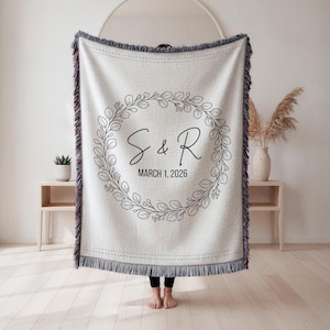 May include: Cream-coloured woven throw blanket with a decorative fringe. The blanket features a circular wreath design with the initials "S & R" and the date "March 1, 2026" in black script. The blanket is displayed indoors.