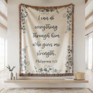 May include: Cream-colored woven throw blanket with the text "I can do everything through him who gives me strength. Philippians 4:13." The blanket has a floral border in shades of blue and a fringed edge.