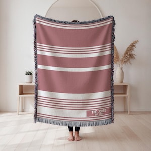 May include: A woven throw blanket with a dusty rose and white striped pattern. The blanket has a fringed edge and the text "THE JONES EST. 2001" in the lower right corner. The blanket is approximately 60 inches wide and 80 inches long.