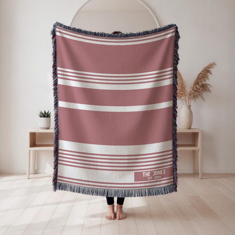 May include: A woven throw blanket with a dusty rose and white striped pattern. The blanket has a fringed edge and the text "THE JONES EST. 2001" in the lower right corner. The blanket is approximately 60 inches wide and 80 inches long.