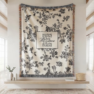 Op de afbeelding: Cr&egrave;mekleurige bloemenwerper met zwart bloemmotief en franjes. De deken heeft de tekst "The Evans" in een zwart omkaderde doos. De deken wordt aan een muur getoond.