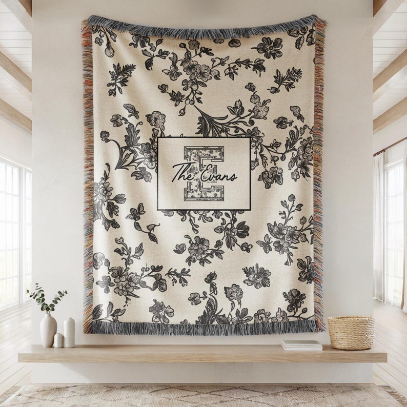 Op de afbeelding: Cr&egrave;mekleurige bloemenwerper met zwart bloemmotief en franjes. De deken heeft de tekst "The Evans" in een zwart omkaderde doos. De deken wordt aan een muur getoond.