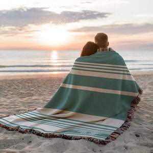May include: A teal and cream striped blanket with fringed edges, draped over two people on a sandy beach. The blanket has horizontal stripes and a personalized inscription. The scene is set at sunset, with the ocean in the background.