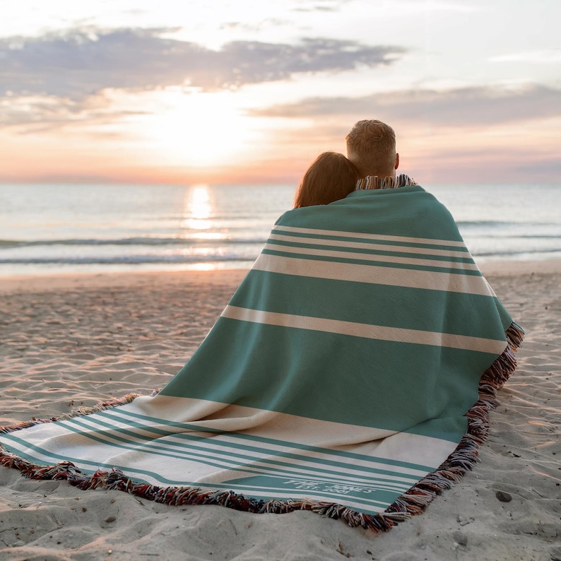 May include: A teal and cream striped blanket with fringed edges, draped over two people on a sandy beach. The blanket has horizontal stripes and a personalized inscription. The scene is set at sunset, with the ocean in the background.