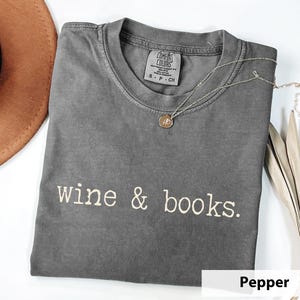 May include: A pepper-colored t-shirt with the words "wine & books" in white script. A silver necklace with a pendant rests on the shirt. A brown hat and dried leaves are in the background.