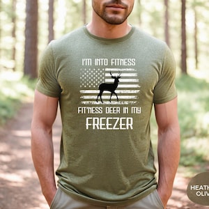 May include: A heather olive green short-sleeve t-shirt featuring a white graphic of an American flag with a black deer silhouette in the center. The text reads I'M INTO FITNESS FIT'NESS DEER IN MY FREEZER in white capital letters.