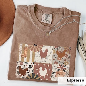 May include: Espresso-colored t-shirt with a patchwork design featuring a cowboy boot, chickens, and floral patterns. The shirt has a crew neck and short sleeves. A silver necklace with a pendant is draped across the shirt.