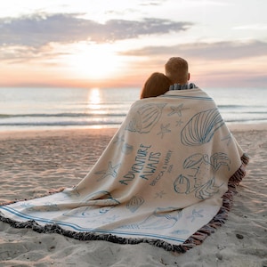 May include: A beige beach blanket with blue seashell and starfish illustrations. The blanket has the words "ADVENTURE AWAITS" and the names "Becca & Harry 2026" printed on it. The blanket is fringed and has blue stripes.