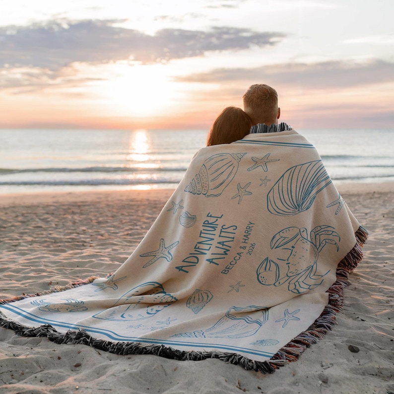 May include: A beige beach blanket with blue seashell and starfish illustrations. The blanket has the words "ADVENTURE AWAITS" and the names "Becca & Harry 2026" printed on it. The blanket is fringed and has blue stripes.