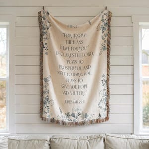May include: Cream-colored woven throw blanket with a floral design in shades of blue and gray. The blanket features the text "FOR I KNOW THE PLANS I HAVE FOR YOU..." from Jeremiah 29:11. The edges have a fringed trim.