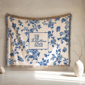 Op de afbeelding: Cr&egrave;mekleurige sprei met een blauw bloemenpatroon en franjes. Het midden heeft een vierkant met de letter "L" en de woorden "The Letteras" in een scriptlettertype. De sprei wordt tegen een neutrale muur getoond.