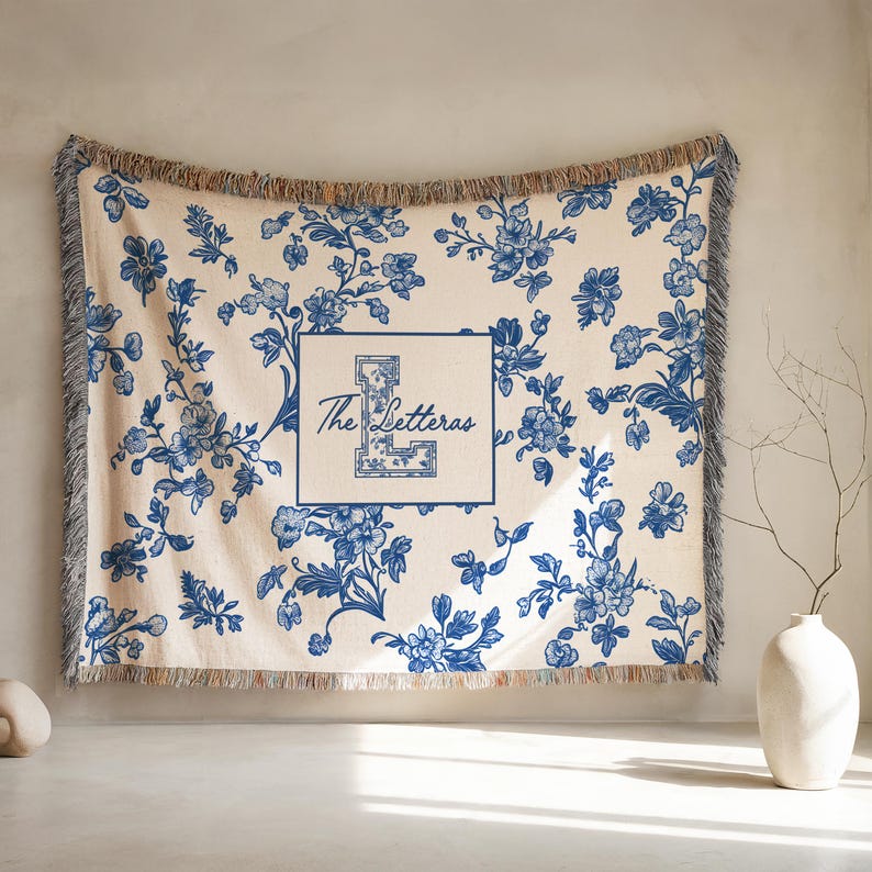 Op de afbeelding: Cr&egrave;mekleurige sprei met een blauw bloemenpatroon en franjes. Het midden heeft een vierkant met de letter "L" en de woorden "The Letteras" in een scriptlettertype. De sprei wordt tegen een neutrale muur getoond.