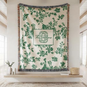 Op de afbeelding: Een cr&egrave;mekleurige sprei met een groen bloemmotief en franjes. De sprei heeft een rechthoekig ontwerp met de tekst "The Derman's" in een groene lijst. De sprei wordt aan een muur getoond.