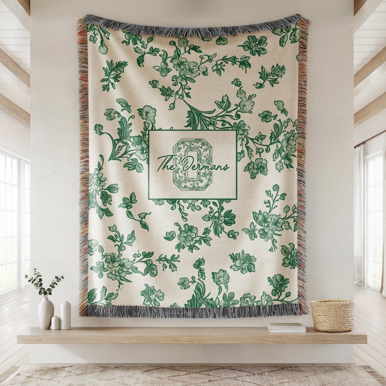 Op de afbeelding: Een cr&egrave;mekleurige sprei met een groen bloemmotief en franjes. De sprei heeft een rechthoekig ontwerp met de tekst "The Derman's" in een groene lijst. De sprei wordt aan een muur getoond.