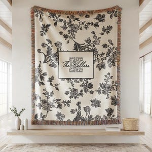 May include: Cream-colored throw blanket with a black floral pattern and fringed edges. The blanket features a rectangular design with the text "The Kellers" in a black frame. The blanket is displayed on a wall.