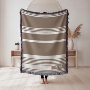 May include: A brown and white striped throw blanket with fringed edges. The blanket has horizontal stripes and a rectangular label with the text "THE JONES EST. 2001". The blanket is displayed indoors.