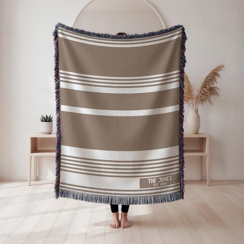 May include: A brown and white striped throw blanket with fringed edges. The blanket has horizontal stripes and a rectangular label with the text "THE JONES EST. 2001". The blanket is displayed indoors.