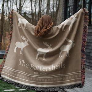 May include: A beige throw blanket with white moose designs and the text "The Battershells est. 2019". The blanket has a decorative border and a fringed edge. The blanket is being held up.