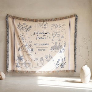 May include: A beige woven throw blanket with a blue illustrated design. The design includes the text "Adventure Awaits!" and various travel-themed illustrations such as a passport, mountains, and a cityscape. The blanket has fringed edges.