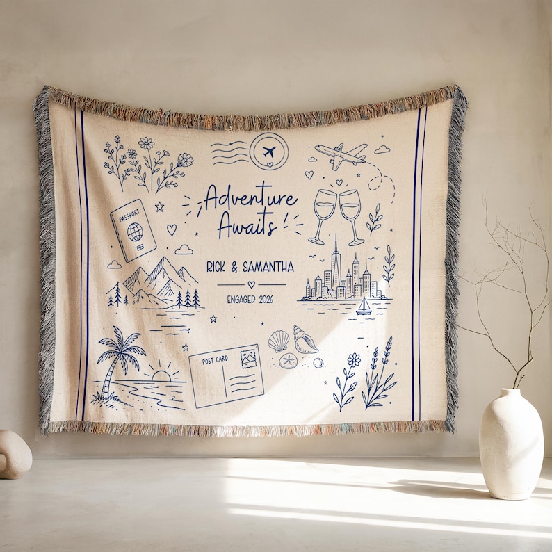 May include: A beige woven throw blanket with a blue illustrated design. The design includes the text "Adventure Awaits!" and various travel-themed illustrations such as a passport, mountains, and a cityscape. The blanket has fringed edges.