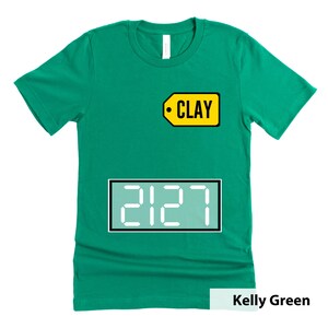 Puede incluir: Camiseta verde Kelly con una etiqueta amarilla que dice "CLAY" y una pantalla digital blanca que muestra "2127".
