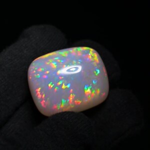 K&ouml;nnte beinhalten: Ein wei&szlig;er Opal-Edelstein mit einem Regenbogen aus Farben, darunter Rot, Orange, Gelb, Gr&uuml;n, Blau und Lila. Der Opal ist im Cabochon-Schliff geschliffen und hat eine glatte, polierte Oberfl&auml;che.