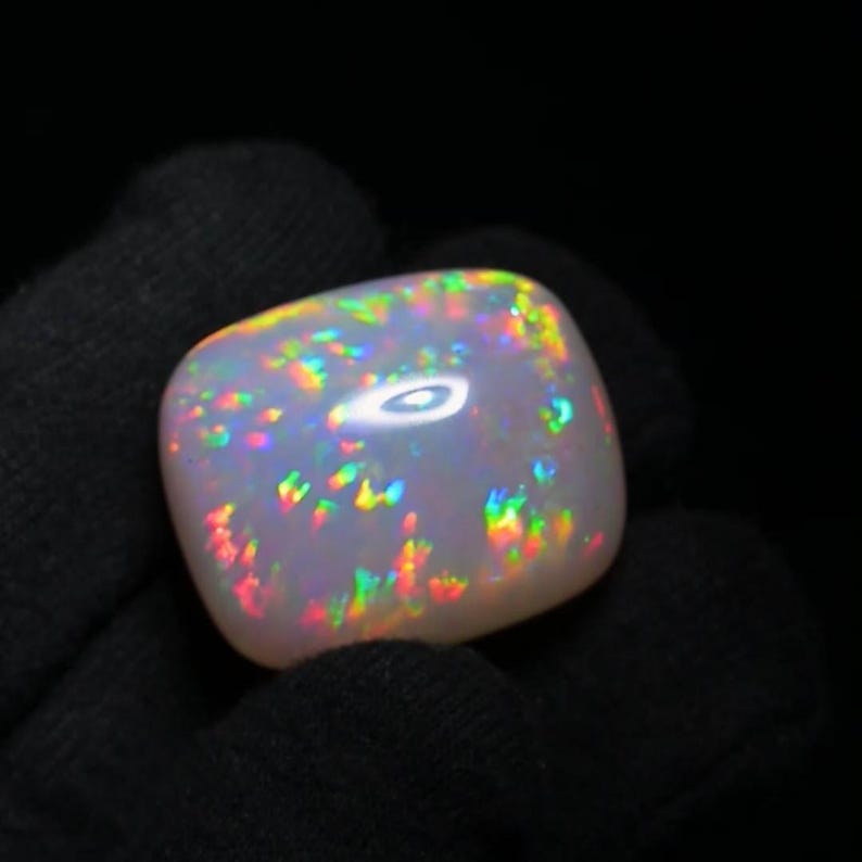 K&ouml;nnte beinhalten: Ein wei&szlig;er Opal-Edelstein mit einem Regenbogen aus Farben, darunter Rot, Orange, Gelb, Gr&uuml;n, Blau und Lila. Der Opal ist im Cabochon-Schliff geschliffen und hat eine glatte, polierte Oberfl&auml;che.