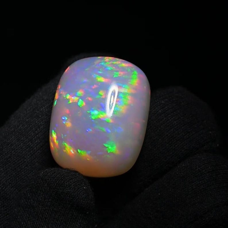 K&ouml;nnte beinhalten: Ein wei&szlig;er Opal-Cabochon mit irisierenden gr&uuml;nen, blauen und rosa Blitzen. Der Opal ist glatt und poliert.