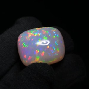 K&ouml;nnte beinhalten: Ein wei&szlig;er Opal-Edelstein mit einem Cabochon-Schliff. Der Opal zeigt einen Regenbogen an Farben, darunter Rot, Orange, Gelb, Gr&uuml;n, Blau und Lila.