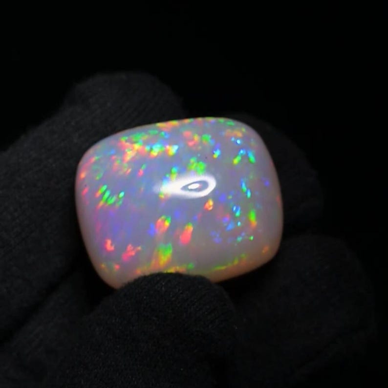K&ouml;nnte beinhalten: Ein wei&szlig;er Opal-Edelstein mit einem Cabochon-Schliff. Der Opal zeigt einen Regenbogen an Farben, darunter Rot, Orange, Gelb, Gr&uuml;n, Blau und Lila.