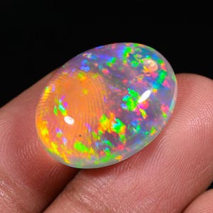 May include: An oval-shaped opal gemstone with a vibrant display of colors. The opal features a translucent body with flashes of green, yellow, orange, and blue. The gemstone is held between fingers against a dark background.