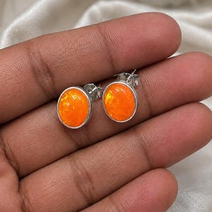 May include: A pair of stud earrings featuring oval-shaped, vibrant orange fire opals set in polished silver-toned metal bezels. Each earring includes a standard post and butterfly clutch backing, showcasing a smooth, rounded cabochon surface.