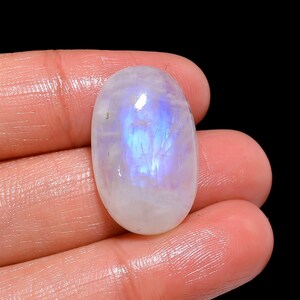 Könnte beinhalten: Ein ovaler Mondstein-Cabochon mit einer durchscheinenden weißen Basis und einer markanten blauen Adulareszenz oder einem Schimmer in der Mitte. Der Edelstein hat eine glatte, polierte Oberfläche und weist sichtbare interne Einschlüsse auf.