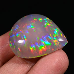 Könnte beinhalten: Ein polierter, birnenförmiger Opal-Edelstein. Der Opal zeigt eine milchig-weiße Grundfarbe mit lebendigen Regenbogenfarben, einschließlich Rot-, Grün- und Blautönen. Der Edelstein ist glatt und hat einen hohen Glanz, vor dunklem Hintergrund.