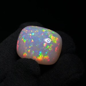K&ouml;nnte beinhalten: Ein wei&szlig;er Opal-Cabochon mit einem Regenbogen aus irisierenden Farben. Der Opal ist ein nat&uuml;rlicher Edelstein mit einer glatten, polierten Oberfl&auml;che.