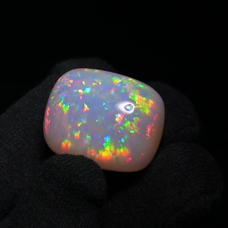 K&ouml;nnte beinhalten: Ein wei&szlig;er Opal-Cabochon mit einem Regenbogen aus irisierenden Farben. Der Opal ist ein nat&uuml;rlicher Edelstein mit einer glatten, polierten Oberfl&auml;che.