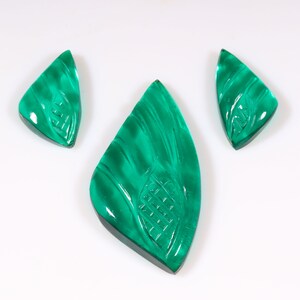 May include: Three green gemstone cabochons with carved designs. The cabochons are cut in a triangular shape with rounded edges.
