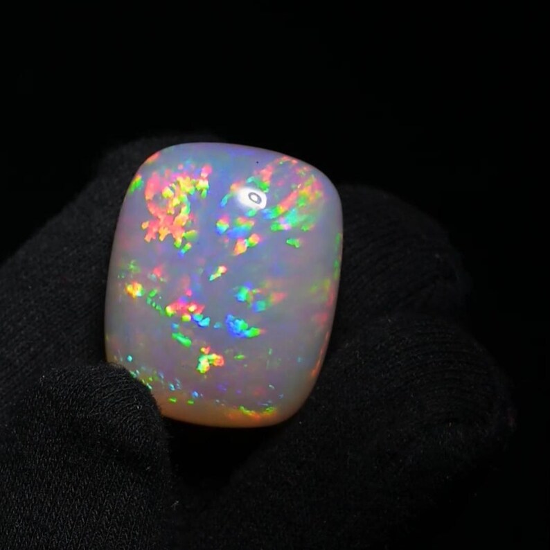 K&ouml;nnte beinhalten: Ein wei&szlig;er Opal-Cabochon mit einem Regenbogen an Farben, darunter Rosa, Orange, Gelb, Gr&uuml;n, Blau und Lila.