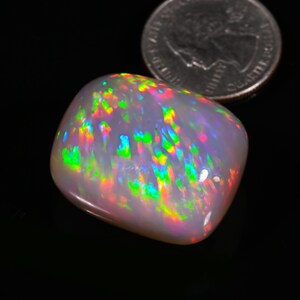 K&ouml;nnte beinhalten: Ein gro&szlig;er, wei&szlig;er, ovaler Opal-Edelstein mit einem Regenbogen aus Farben, darunter Gr&uuml;n, Blau, Rot und Orange. Der Opal liegt auf einer schwarzen Oberfl&auml;che.