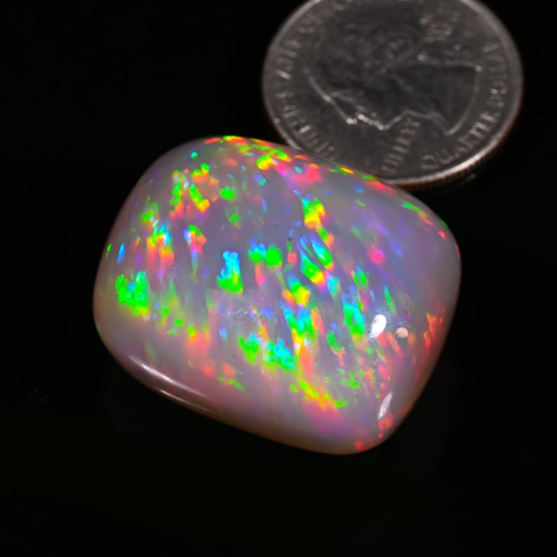 K&ouml;nnte beinhalten: Ein gro&szlig;er, wei&szlig;er, ovaler Opal-Edelstein mit einem Regenbogen aus Farben, darunter Gr&uuml;n, Blau, Rot und Orange. Der Opal liegt auf einer schwarzen Oberfl&auml;che.